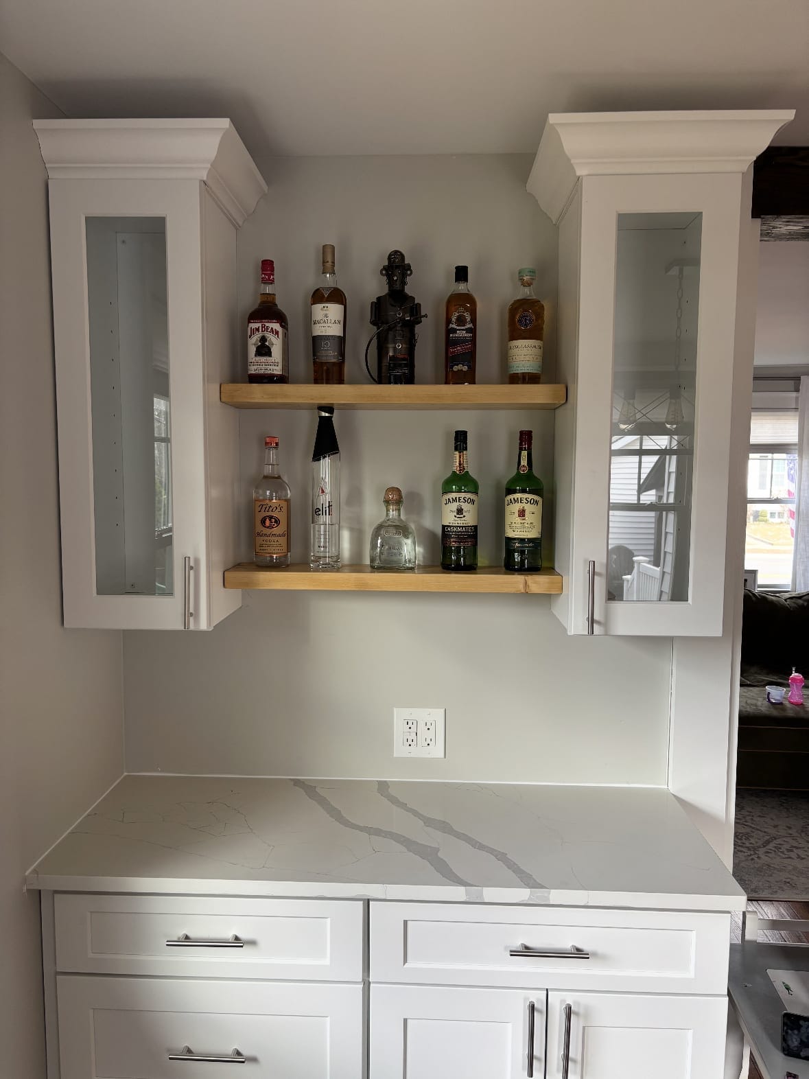 Two wood floating shelves holding bottles between white glass-front cabinets